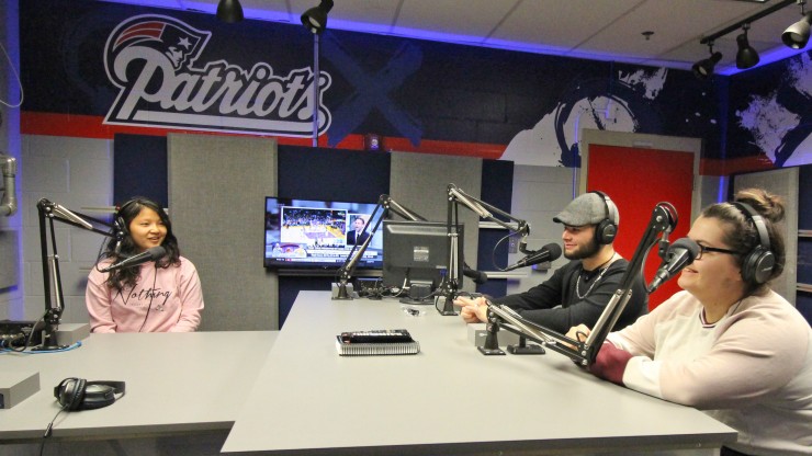 Three students inside the Patriots radio broadcasting studio, receiving hands-on experience at the radio broadcasting microphone. 