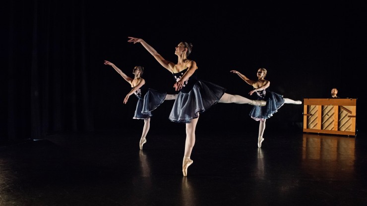 Three ballet students on their toes during a pointe dance onstage with a pianist playing in the background. 