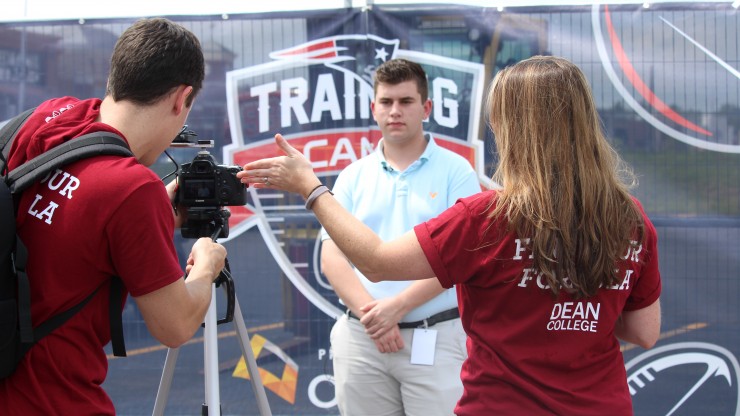 A student stands in front of a camera as two other people work the camera equipment while broadcasting from Patriots Training Camp at Gillette Stadium. 