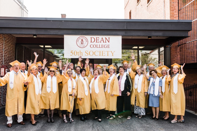 image of alumni from the 50th society wearing graduation caps and gowns smiling in front of the dean college 50th society sign.