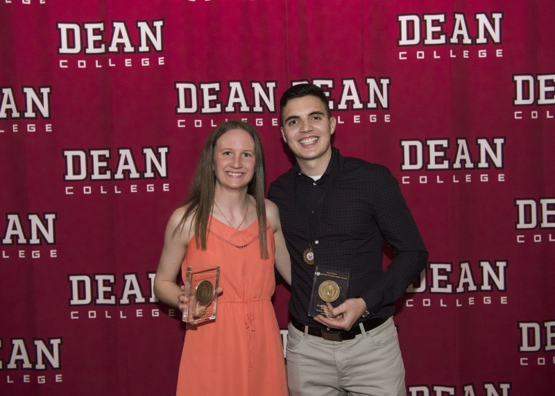 Image of two students holding awards for athletic and academic achievement and standing in front of a dean college backdrop