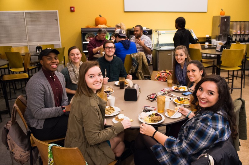 image of students sitting around a dinner table in the dining center smiling at the camera with their plates full of food from thanksgiving dinner.